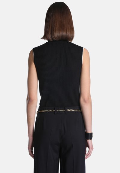 Femme aux cheveux bruns et lisses, portant un haut noir côtelé sans manches, un pantalon noir avec une ceinture chaîne dorée et un bracelet manchette noir, vue de dos.