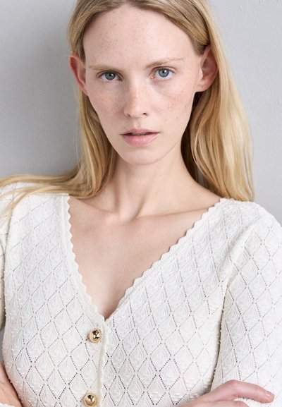 Witte, gestructureerde cardigan met een ruitpatroon, voorzien van een V-hals en golfranden, versierd met twee gouden knopen in het midden.