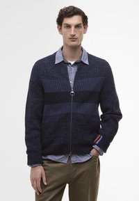 Marineblauwe gebreide cardigan met een rits aan de voorkant, gestructureerde strepen en een kleurrijke accent op de manchet, gedragen over een lichtblauw shirt.