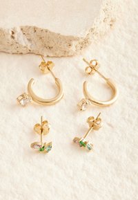 Zwei Paar goldene Ohrstecker auf cremefarbener Stoffunterlage; ein Paar mit klaren Steinen an Halbkreolen, das andere mit drei kleinen farbigen Edelsteinen.