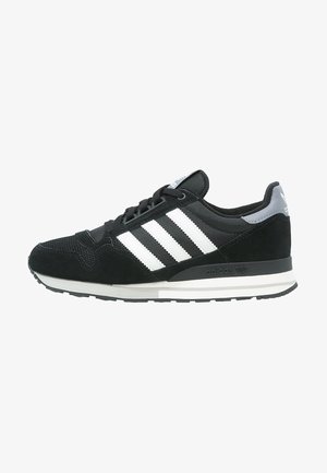 Baskets Adidas noires avec des bandes blanches, languette grise au talon, semelle noire et fermeture à lacets, vues de côté gauche.