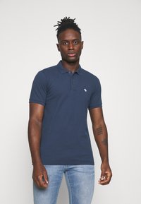 Polo shirt blu navy in cotone, con colletto a tre bottoni e un piccolo logo ricamato sul petto. Indossato con jeans azzurri chiari.