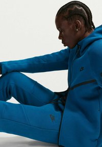 Μπλε φόρμα Nike από μαλακό υλικό, με φούτερ με φερμουάρ και μαύρες λεπτομέρειες και λογότυπα, απαλή υφή και εφαρμοστή σχεδίαση.