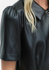 Zwarte leren blouse met korte puffmouwtjes, een puntige kraag en een ronde zilveren sluiting bij de hals. Gladde textuur.