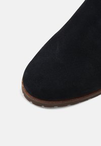 Botte en daim noir avec un bout pointu, dotée d'une semelle en caoutchouc brun contrastante et d'une tige texturée. Aucun élément métallique visible ni accent supplémentaire.