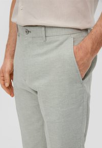 Gros plan sur une personne portant un pantalon à carreaux gris clair avec une main dans la poche et une chemise beige rentrée.