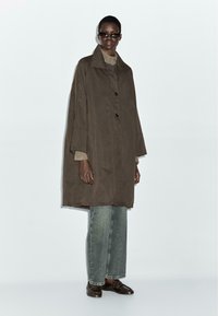 Cappotto oversized marrone con colletto rigido, chiusura a bottoni e maniche arrotolate, abbinato a jeans in denim chiaro e scarpe marroni. Tessuto dalla texture liscia.