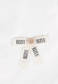 Witte stof versierd met een lichtbeige lint met "BIZUU" in zwart gedrukt. Het lint is in een strik om een houten knop gestrikt.