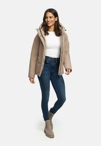 Beige gewatteerde jas met capuchon, doorgestikte ontwerp, rits aan de voorkant en zijzakken, gecombineerd met strakke donkerblauwe jeans en beige enkellaarsjes.