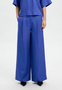 Mėlynos plačios kelnės iš lengvos medžiagos, su raukšlėmis ir lygiu paviršiumi, švariu užbaigimu ir priglundančiu liemeniu.