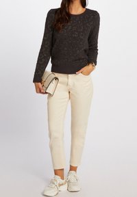 Femme portant un pull sombre à motifs, un pantalon beige taille haute, des baskets blanches, tenant un sac à main crème et portant un bracelet en or sur fond blanc.