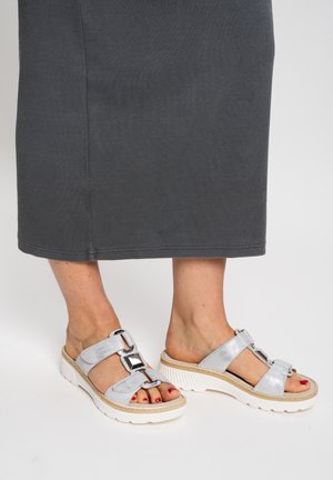 Silberne Sandalen mit offenem Zehenbereich und dekorativen Schnallenriemen, getragen an Füßen mit rot lackierten Fußnägeln, kombiniert mit einem dunkelgrauen Rock oder Kleid.