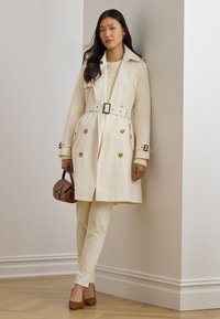 Cream trench coat with double-breasted buttons, belted waist, and side pockets, paired with cream pants and brown handbag.