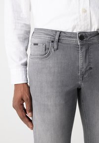 Vaqueros de mezclilla gris con un ajuste slim, que cuentan con una cintura de altura media, bolsillos frontales y un sutil detalle de marca en el bolsillo trasero.