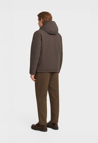 Giubbotto imbottito marrone con cappuccio, caratterizzato da una finitura liscia e opaca. Abbinato a pantaloni marroni e scarpe scure, visto da dietro.