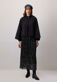 Woman wearing a blue beret, black cropped jacket with wide sleeves, long pleated black and white skirt, and black leather ankle boots, standing.