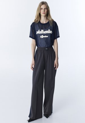 Vrouw staand, gekleed in een marineblauw doorschijnend T-shirt met de tekst "Authentic" en wijde grijze broek met zwarte puntige schoenen tegen een witte achtergrond.