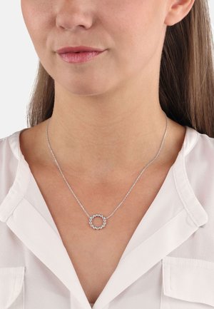 Collar de plata con un colgante circular adornado con pequeñas piedras preciosas, suspendido de una cadena delicada, llevado sobre una blusa clara.