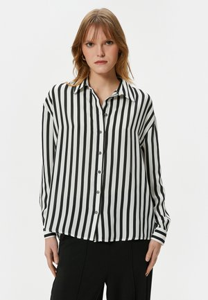 Camisa de botones a rayas blancas y negras con cuello, mangas largas y dobladillo redondeado. Presenta una tela suave y ligera y botones visibles.