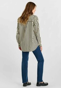 Langsleeve shirt met verticale zwarte en crème strepen, losse fit, licht gebogen zoom. Gecombineerd met donkerblauwe jeans en zwarte schoenen.