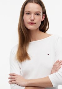 Giovane donna con capelli lunghi e lisci indossa un top bianco con un piccolo logo, braccia incrociate, guarda con calma la fotocamera su sfondo bianco.