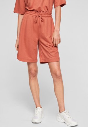 Personne portant un short corail à cordon et un haut assorti, debout en baskets blanches sur un fond gris uni.