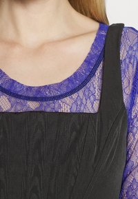 Černé oblečení s texturovaným vzorem, charakterizované vrstveným fialovým krajkovým topem se zvlněným výstřihem a kontrastními tmavými ramínky.