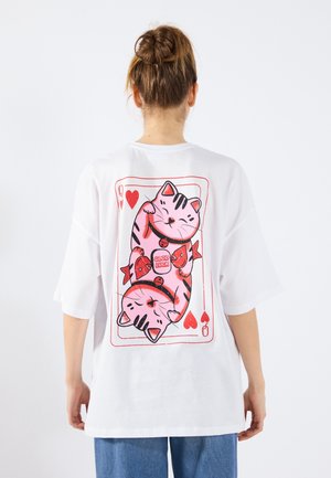 Persoon draagt een wit T-shirt met een groot speelkaartontwerp van twee roze katten die vissen vasthouden en een "Good Luck" label op de achterkant.