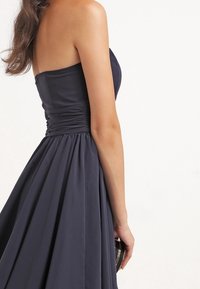 Marinblå, ärmlös klänning med en åtsittande liv och samlat midjeband, som flyter ut i en full kjol. Slät tyg, minimala detaljer.