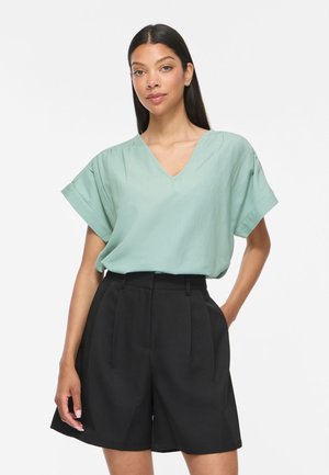 Femme portant un chemisier vert clair à col en V avec des manches courtes et un short plissé noir taille haute, debout avec une main dans la poche.
