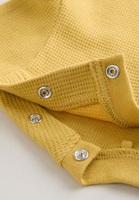 Tissu en maille gaufrée jaune avec trois boutons-pression argentés sur les bords pliés, montrant la texture et les détails de la fixation.