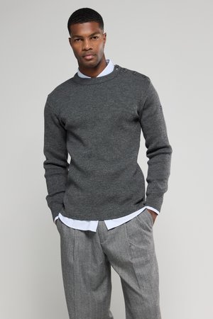 Pull en tricot gris avec un col rond et des détails de boutons sur l'épaule, superposé à une chemise à col bleu clair, associé à un pantalon à rayures.