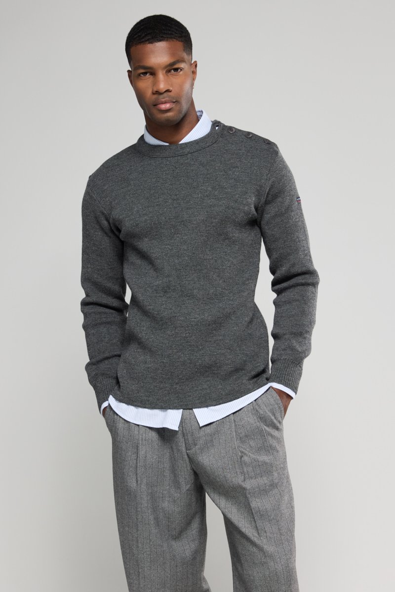 Pull en tricot gris avec un col rond et des détails de boutons sur l'épaule, superposé à une chemise à col bleu clair, associé à un pantalon à rayures.