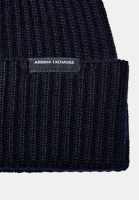 Berretto lavorato a maglia blu navy con una texture a coste e un'etichetta nera che riporta "ARMANI EXCHANGE" in bianco. Tessuto morbido e stretch.