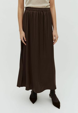 Kobieta ubrana w ciemnobrązową maxispódnicę z bocznym rozcięciem, w połączeniu z ciemnymi szpiczastymi butami i czarnymi skarpetkami, na jednolitym tle.