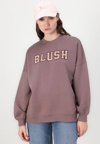 Smešak smeđi oversized dukserica s natpisom "BLUSH" u krem boji; rebrasti manžeti i rub. Uparen s laganom ružičastom kapom s dekorativnim tekstom.