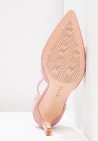 Zapato de tacón alto de ante rosa con punta afilada, tira en el tobillo y suela nude texturizada con líneas onduladas y nombre de la marca en relieve.