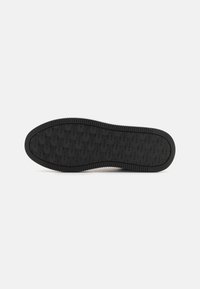 Semelle de chaussure en caoutchouc noir avec motif de grip texturé et logos du nom de la marque sur toute la partie inférieure, présentée sur un fond blanc uni.