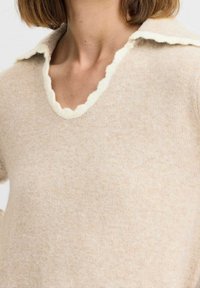 Pull beige avec un col en V festonné bordé de crème. Tissu doux avec une finition légèrement texturée. Manches longues, coupe décontractée.