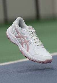 Scarpa da ginnastica bianca con accenti rosa, materiale sintetico, punta arrotondata, lati in rete e suola testurizzata. Allacciatura frontale e marchio ASICS visibile.