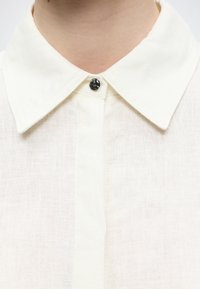 Κοντινό πλάνο ατόμου που φοράει λευκό πουκάμισο με μυτερό γιακά που κλείνει με ένα μαύρο κουμπί στο λαιμό.