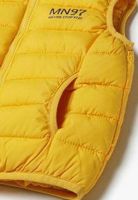 Gelber Puffer-Weste mit diagonaler Steppung, Reißverschluss und seitlichen Taschen. Mit der blauen Stickerei "MN97 NEVER STOP 91" auf der Vorderseite.