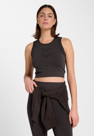 Kobieta w czarnym bezrękawniku typu crop top i czarnych legginsach, z przewiązaną wokół talii bluzą, stojąca na tle jednolitej białej ściany.