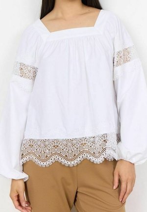 Femme portant un chemisier blanc à manches longues avec des inserts en dentelle sur les manches et l'ourlet, assorti à un pantalon marron clair.