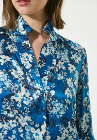 Blusa di satin con sfondo blu e motivi floreali in bianco, azzurro chiaro e blu navy scuro. Design con colletto alto di rilievo.