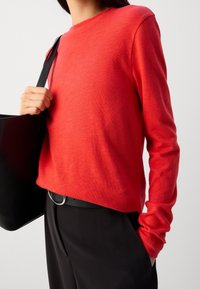 Maglione rosso a maniche lunghe realizzato in un materiale morbido, con scollo rotondo, design aderente, abbinato a una borsa nera e a una cintura nera con fibbia circolare.