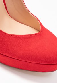 Chaussure à talon haut en daim rouge avec un décolleté bas, semelle compensée et doublure intérieure lisse, présentant un bout arrondi et des détails de couture.