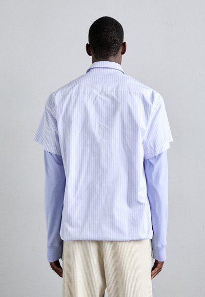 Camicia a maniche corte di colore azzurro chiaro con righe verticali, indossata sopra una camicia a maniche lunghe di colore lavanda. Tessuto morbido, vestibilità rilassata.