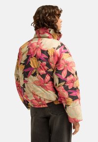 Personne aux cheveux bouclés debout de profil, portant une doudoune colorée à motifs floraux et un jean foncé sur un fond uni.