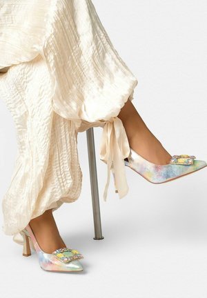 Pies con tacones altos multicolor pastel con hebillas joya, combinados con pantalones fluidos color crema con textura atados en los tobillos.